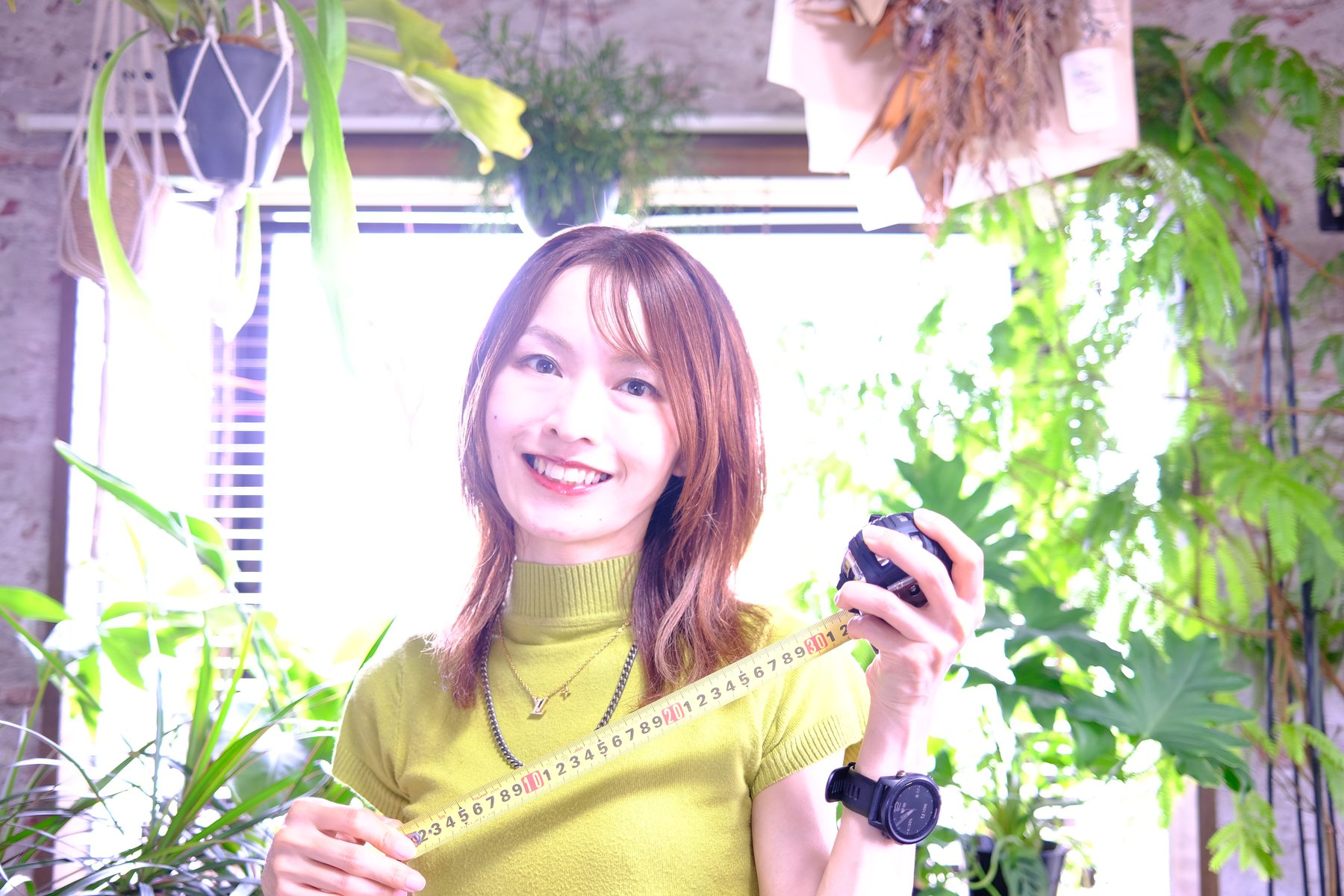 植物に囲まれた明るい室内で、メジャーを手に微笑む代表
